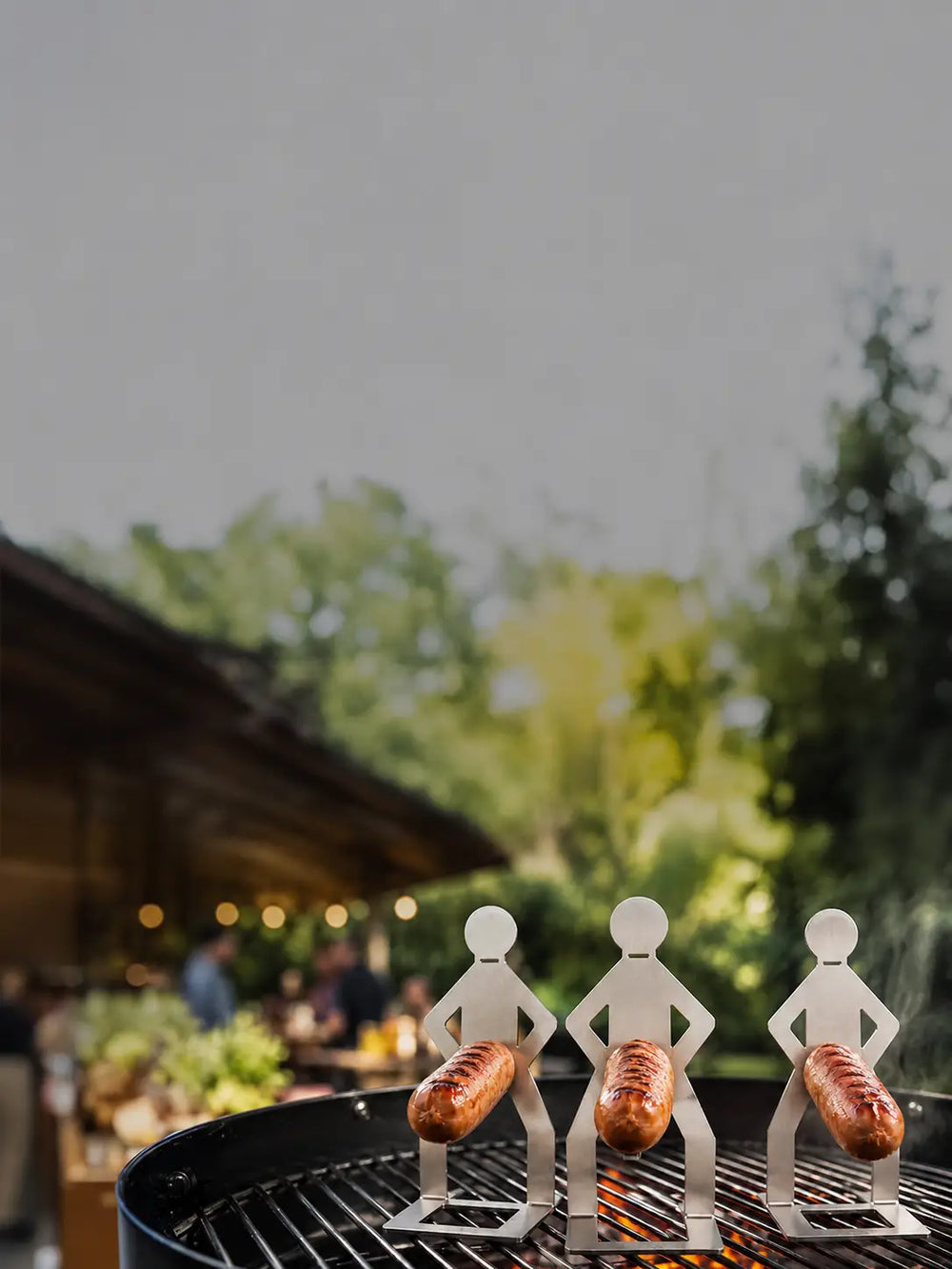 MeatRider Bratwursthalter 3er-Set aus Edelstahl mit gegrillten Bratwürsten auf dem Grill als praktisches BBQ-Zubehör für gleichmäßiges Grillen.