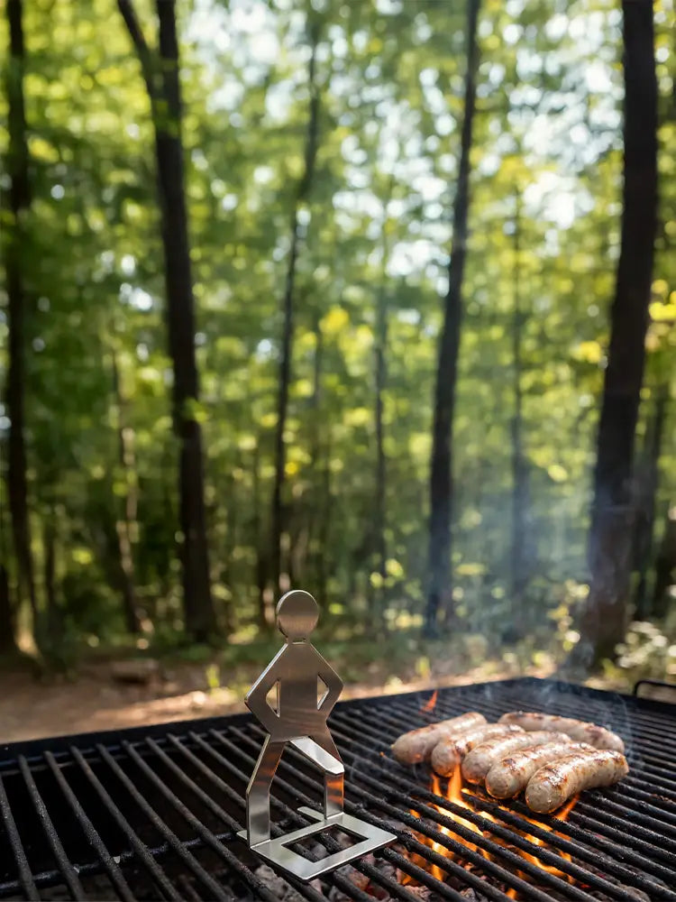 MeatRider Bratwursthalter aus rostfreiem Edelstahl auf einem Grillrost in natürlicher Waldkulisse – hochwertiges Grillzubehör für perfekt gegrillte Bratwürste und echtes Outdoor-BBQ-Erlebnis.